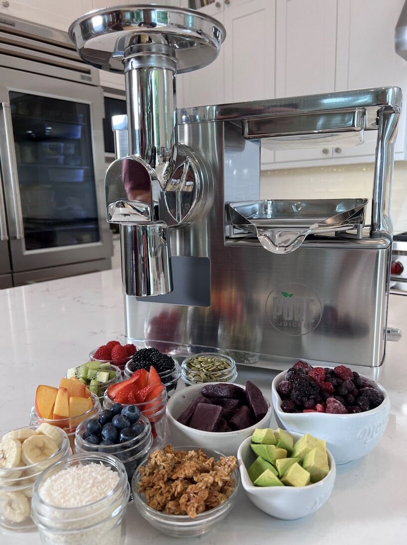 A juicer on a kitchen counter with bowls of berries, avocado, bananas, granola, and other açaí bowl toppings