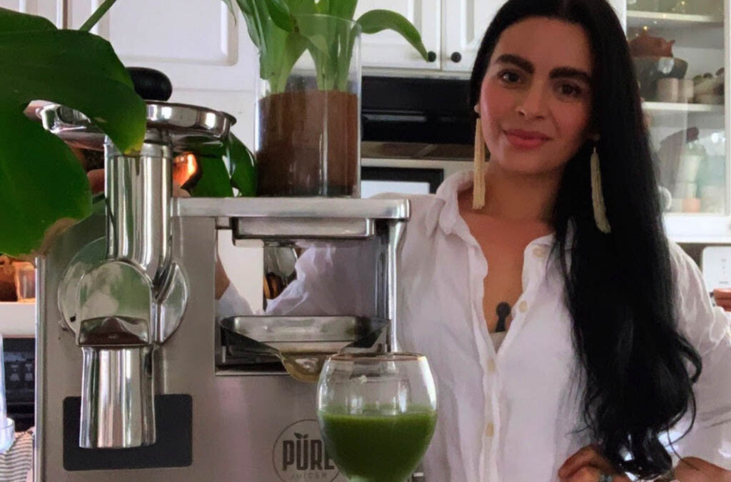 Bekah Sheehan standing next to a PURE Juicer holding a glass of green juice