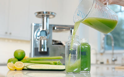 Green juice poured from a measuring pitcher into bottles beside lemons, cucumber and celery