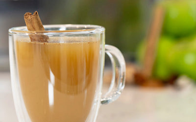 A glass mug full of warm, mulled apple cider, garnished with a cinnamon stick, sits on a countertop
