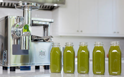 Five bottles of green juice lined up on a kitchen counter next to a PURE Juicer