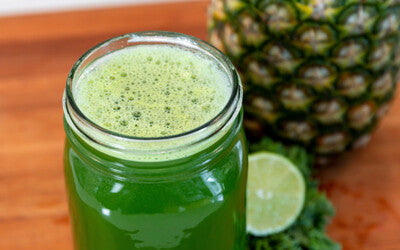 A jar of green juice with a lime wedge, kale and half a pineapple