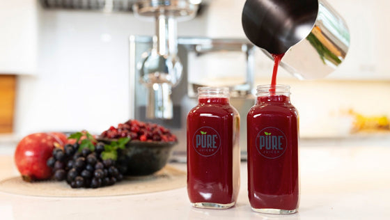Deep red berry juice poured from a steel pitcher into two bottles, with grapes and pomegranate nearby
