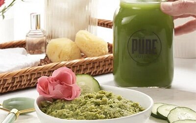 A white bowl of creamy green cucumber face mask made from juice pulp, with a jar of cucumber juice behind it