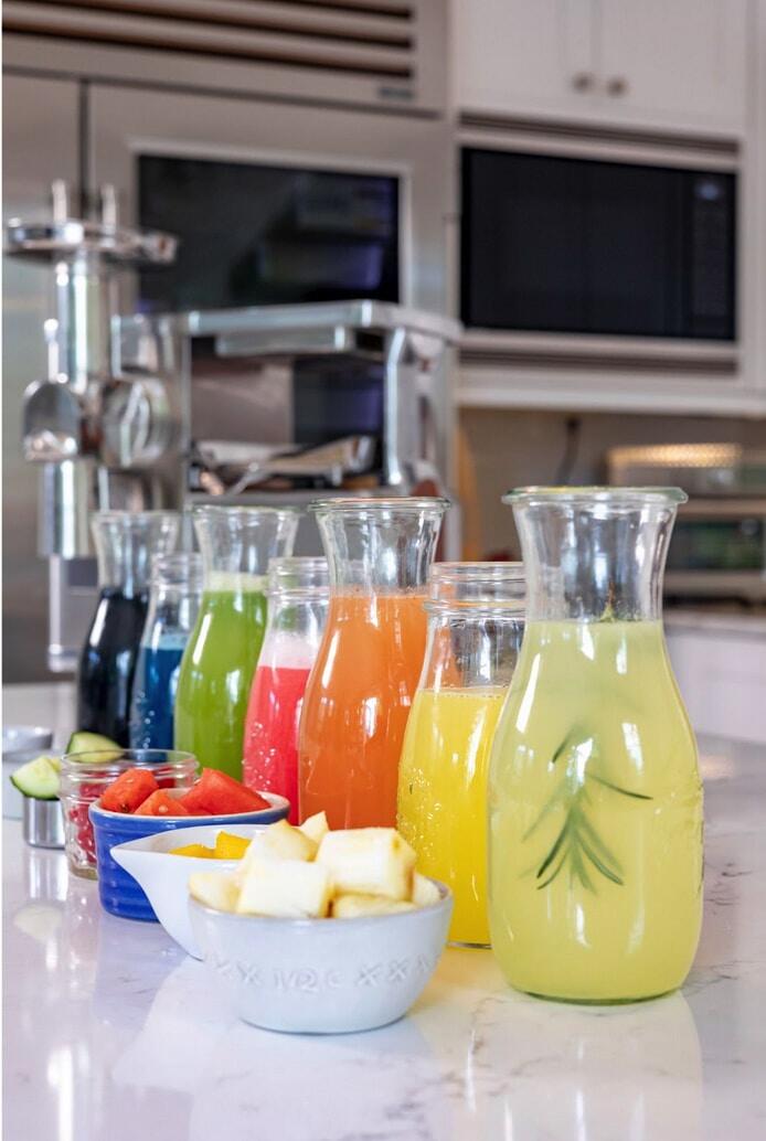 A row of carafes filled with lemonades in yellow, orange, pink, green, blue and black, with bowls of cut fruit in front of a juicer