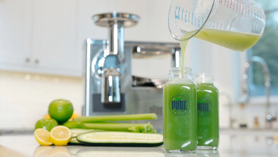 Green juice being poured into bottles in front of a PURE Juicer, with celery, cucumber, lemon and lime