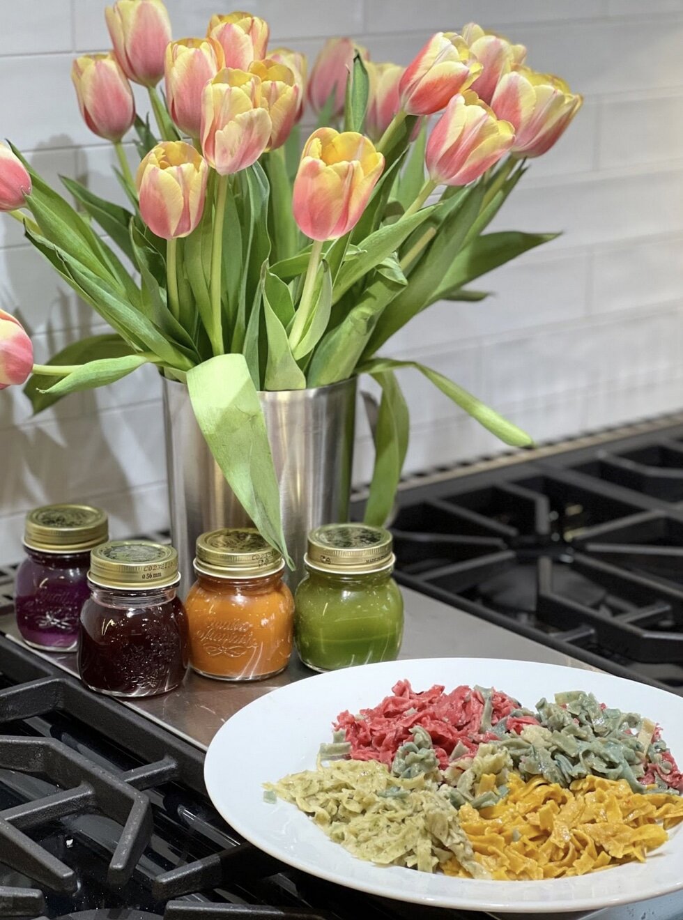 Plate of colorful pasta beside jars of beet, carrot and green juice and a vase of tulips