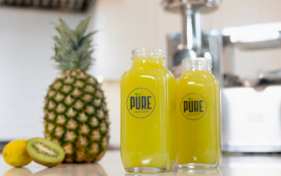 Two bottles of yellow juice next to a pineapple, kiwi and lemon