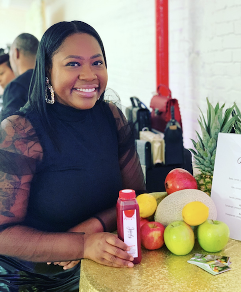 Markia McClenton of Juiced By Ke holding a bottle of cold-pressed juice beside a display of fruit